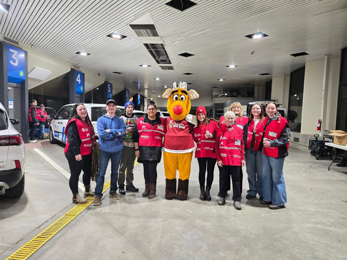 A line-up of nine Operation Red Nose volunteers in red team jackets, standing with Rudy in the vehicle bays they work out of.
